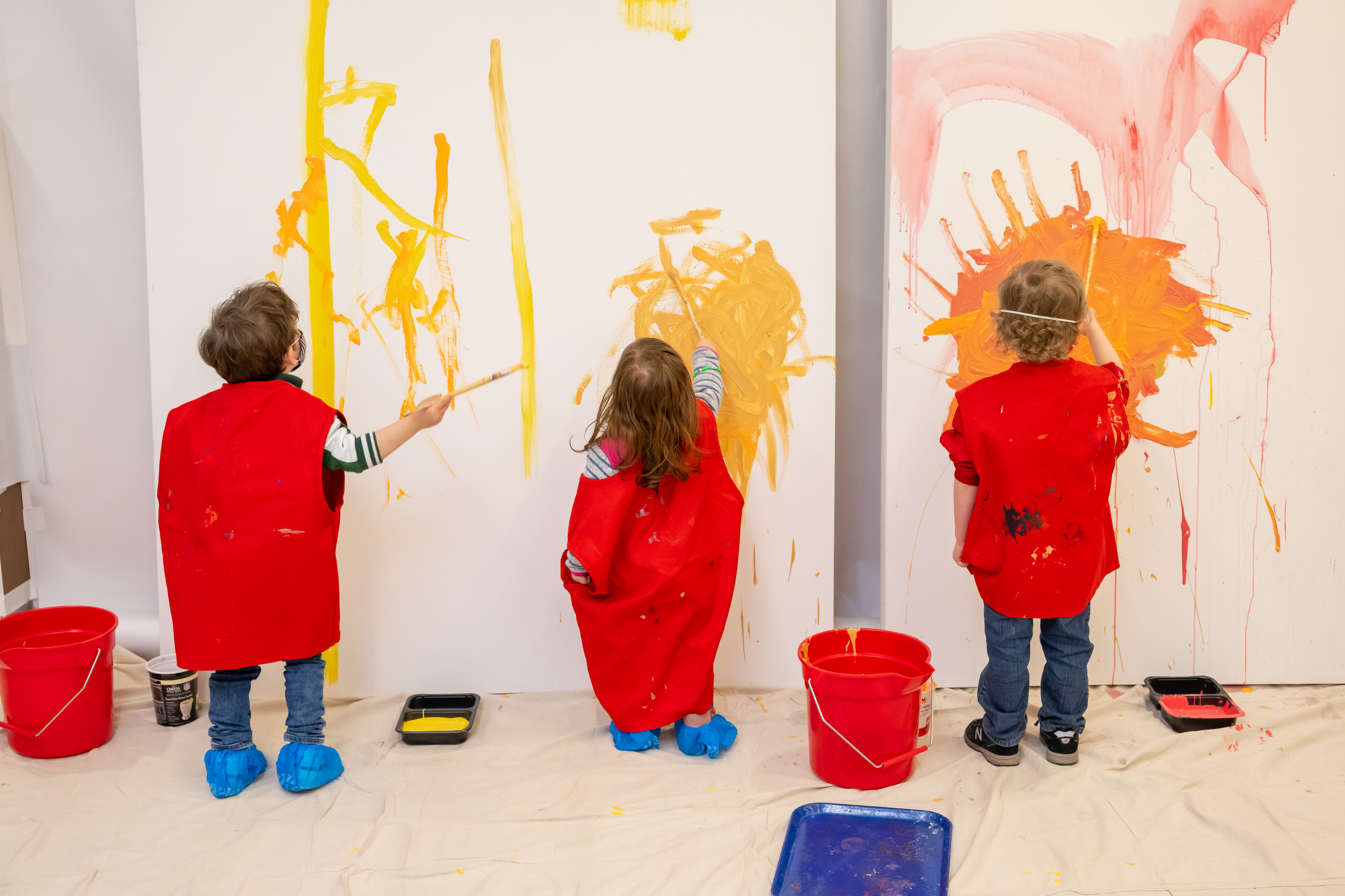 Three children in art smocks splatter orange, yellow, and red paint on three large canvases.