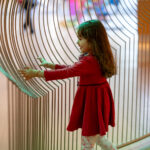 A little girl smiles while she feels a wire sculpture of Mary Flanagan’s Topophilia (Tunnel) and Topophilia (Hill) inside the Joseph Education Center at the Baltimore Museum of Art