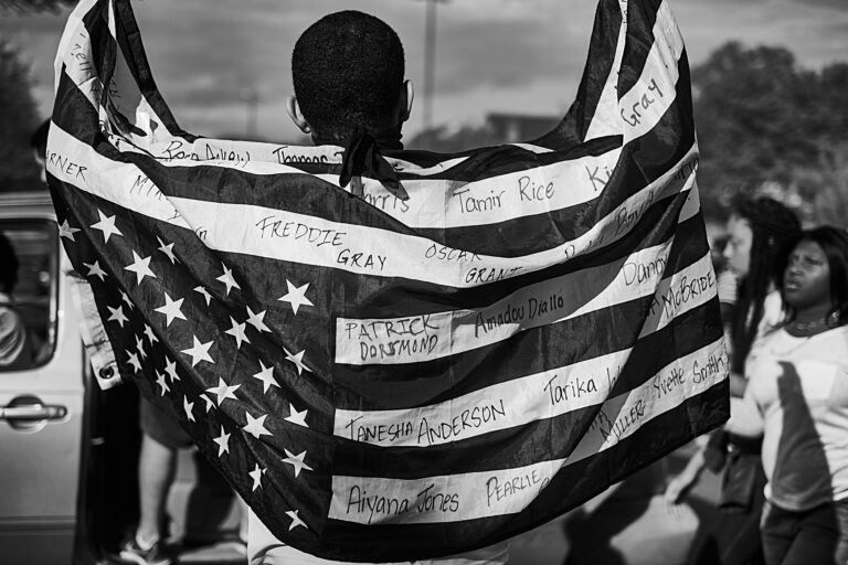 A black and white photo of a person holding an upside down American flag with names written in the white stripes of victims of police brutality. Photographed by Devin Allen in Baltimore during the 2015 Baltimore Uprising