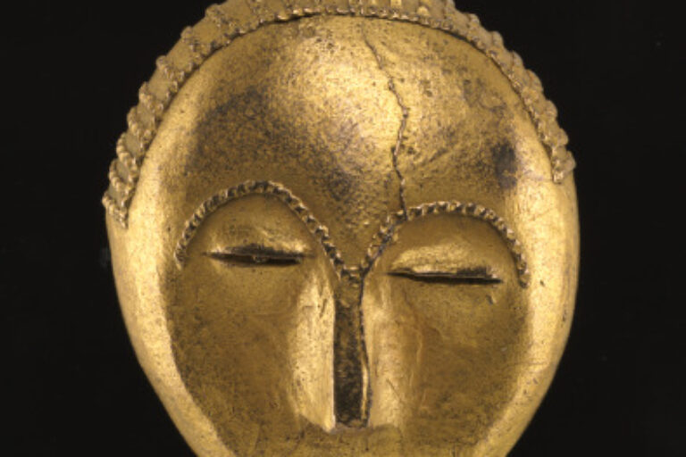 A miniature gold-alloy mask with closed eyes, a narrow nose, and a subtle smile. The top edge of the mask is textured with a pattern resembling braided hair.