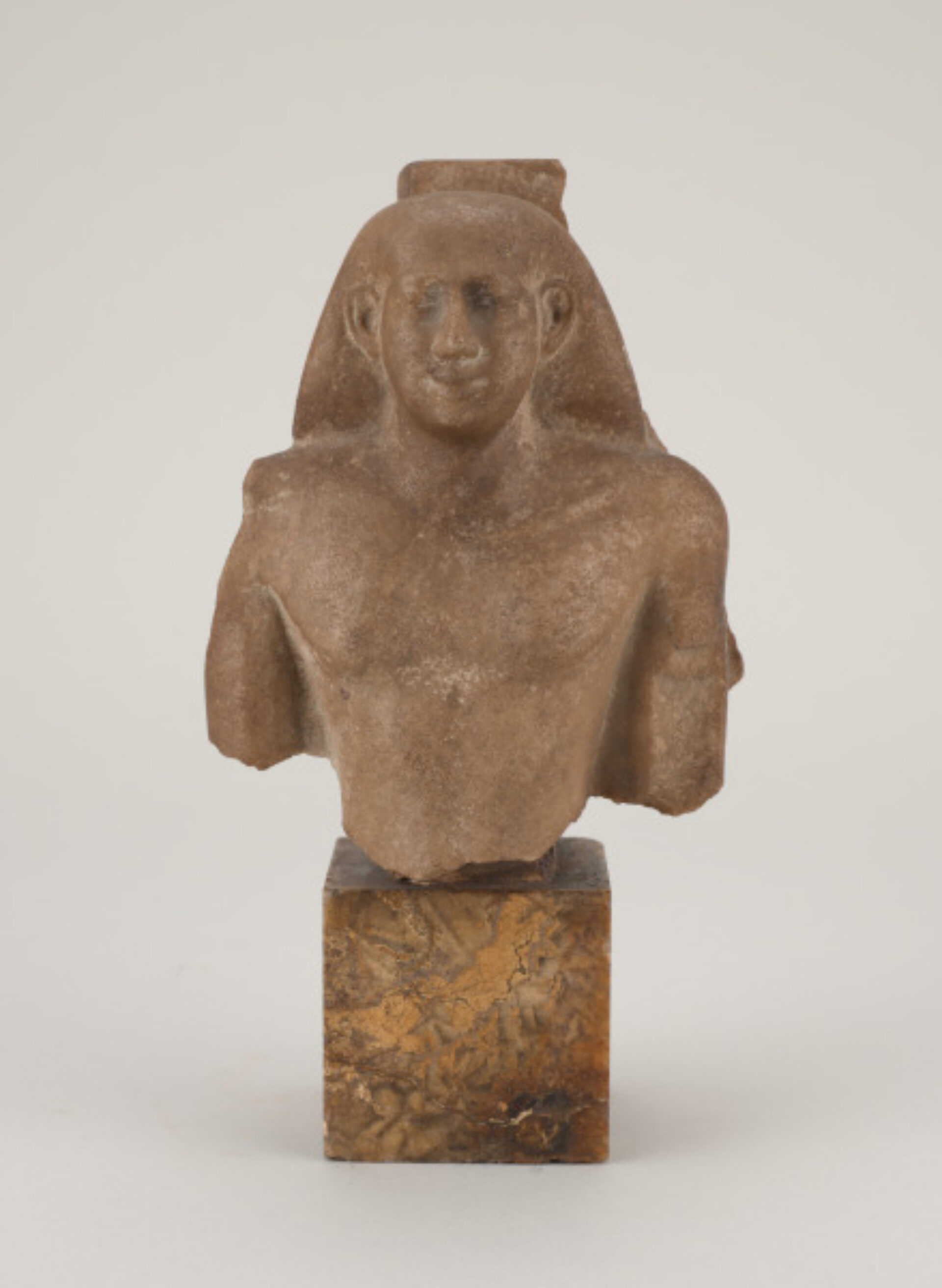 A small stone bust of an ancient Egyptian figure with a headdress typical of Egyptian royalty mounted on a cube of marble-like stone.