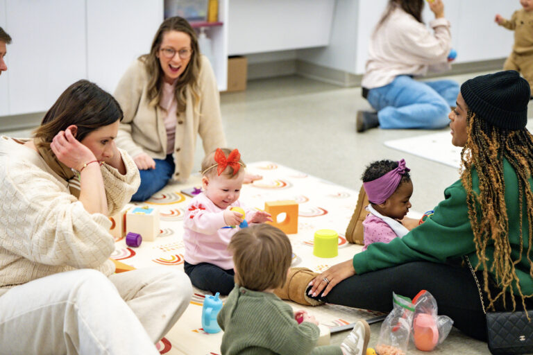 Three smiling caretakers sit on a blanket with three babies playing with colorful toys at the Baltimore Museum of Art's Baby Art Date event in February of 2026.