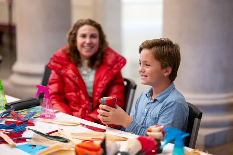 A smiling child holds a colorful string craft while an adult smiles while sitting with him at a table full of supplies at the Baltimore Museum of Art Community Opening in 2023.