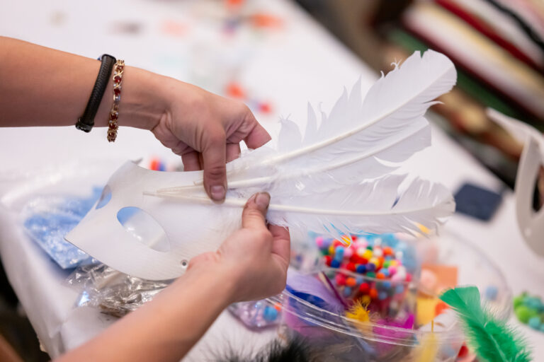 A closeup shot of hands holding together the pieces of a white mask with white feathers.